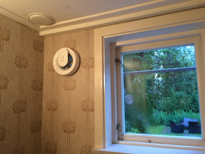 Pax Calima ventilationskontroll monterad på vägg intill ett fönster med utsikt över en trädgård, snart ansluten via utanpåliggande kabel.