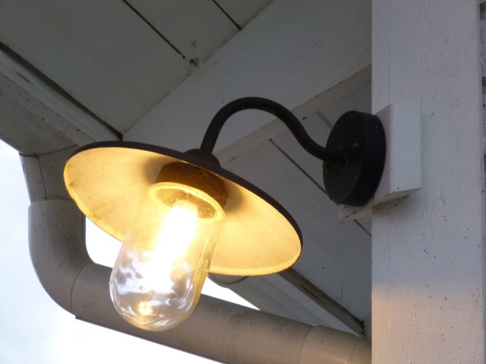 Uppvinklad bild av en utomhuslampa som är installerad under ett takutskjut, med tänd glödlampa och synliga kablar.