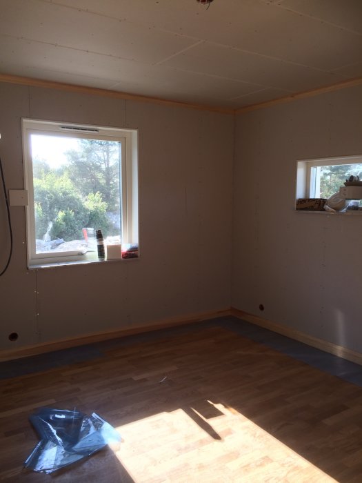 Ett ofärdigt rum med nakna gipsskivor på väggarna, trägolv och ett fönster som släpper in solljus.