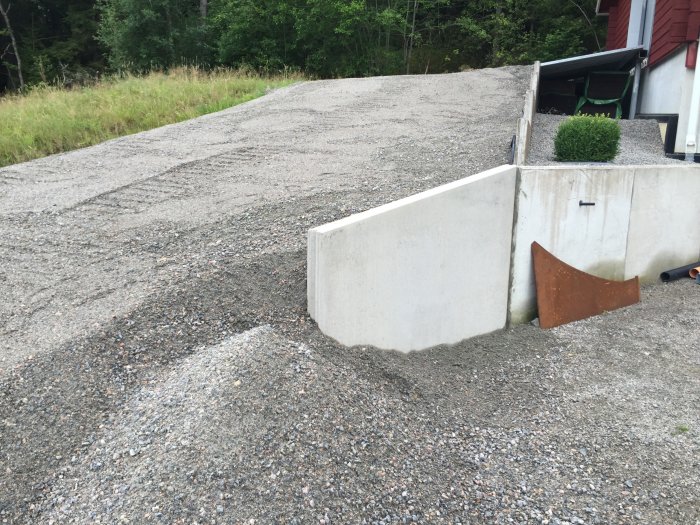 Grusuppfart med nylagd betongstödstruktur bredvid garage och en mur mot skogen.