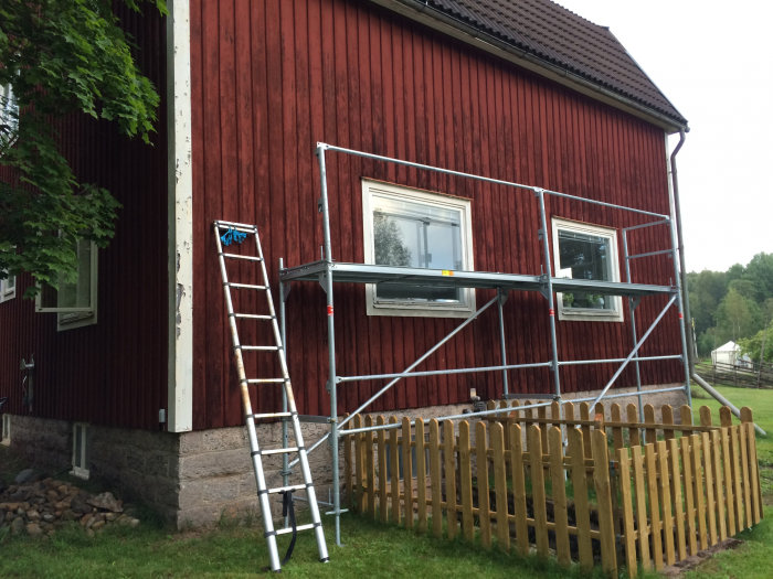 Byggställning monterad vid röd trähusfasad, med stege och ofärdigt räcke.