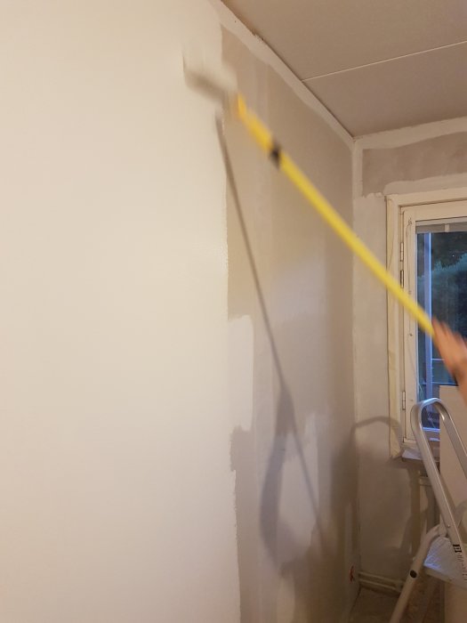 Målning av vägg i "Lammull" nyans S1000-N, roller mitt i draget över tidigare gipsvägg, nära fönster.