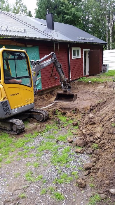 En grävmaskin gräver för tillbyggnad vid ett rött hus med svart tak.