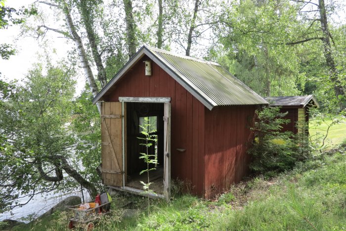 Renoverad röd bod med öppen dörr och nytt tak vid vattnet omgiven av grönska och träd.