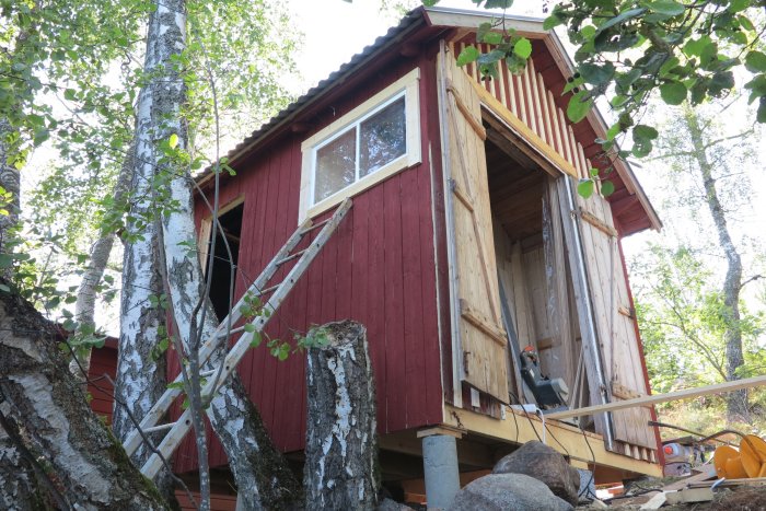 Renoverad bod med nytt bjälklag och delvis ny panel, stödd av temporär ställning bland träd och stenar.