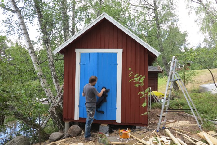 Person renoverar en röd stuga med blå dörr, material och stege synliga.