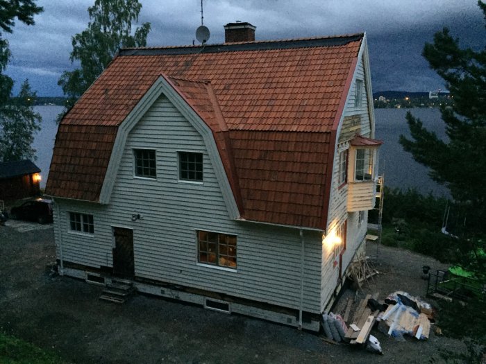 Kvällsbild av ett renoveringsprojekt på ett hus med rött tak och skorsten vid sjö, byggnadsmaterial på marken.