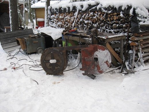 Svetsutrustning och arbetsverktyg på snöbelagd mark framför vedstapel.