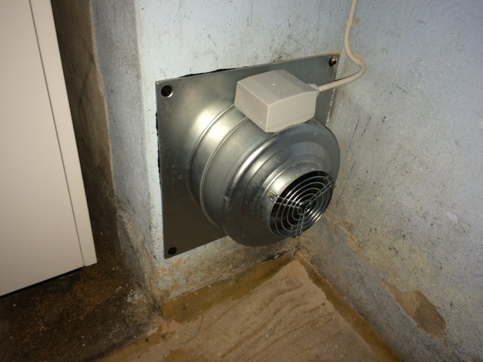 Fläkt installerad i vägg med provisorisk kabeldragning och synlig betong.