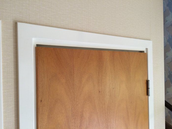 En träinnerdörr med nerkapad nederkant för ventilation, mot en vit dörrkarm och tapetserad vägg.