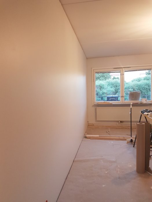 Nymålat vardagsrum med släta väggar och en fönstervägg, osatta socklar och byggmaterial på golvet.