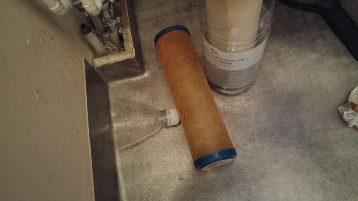 Brunfärgat vattenfilter liggandes på en grå yta bredvid en tom plastflaska och ett rent filter.