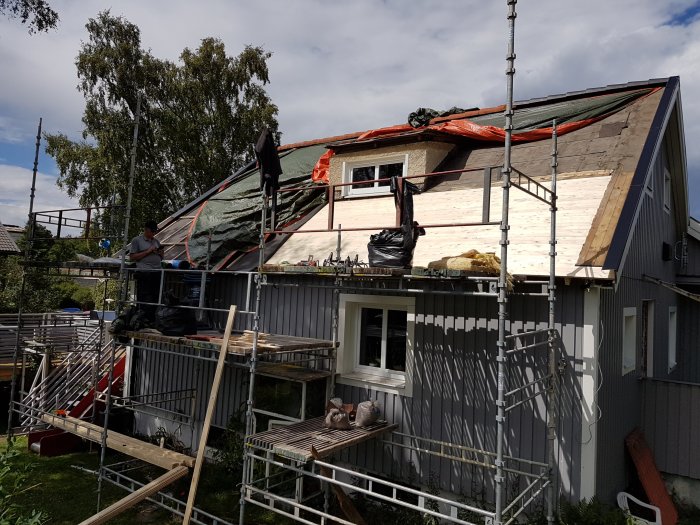 Ställningar runt en halvfärdig husfasad under renovering, med isolering och träbeklädnad synlig.