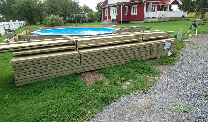 Leverans av byggmaterial från bygghandel, staplade brädor vid en grusgång, röd villa och pool i bakgrunden.