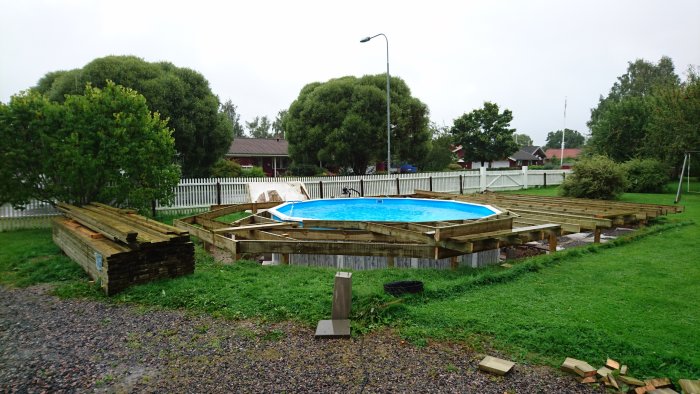 Byggprojekt av trädäck runt ovanmarkspool med leverans av byggmaterial på en gråmulen dag.