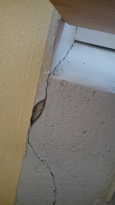 Sprukket vegg med avflassende grå maling rundt vinduskarm, viser behov for restaurering og spørsmålet om riktig verktøy og spakkelarbeid.