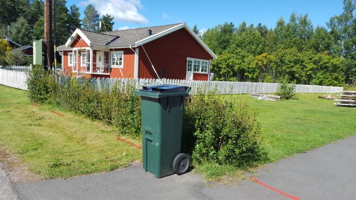 Röd markering på gräsmatta som visar var elledningar kan ligga nära ett rött hus med vit staket och gröna buskar.