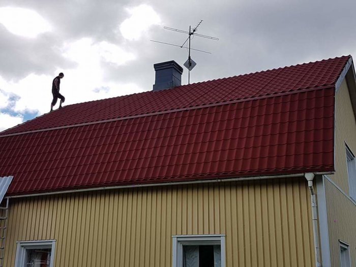 Person som går på nylagt rött tak på ett gult hus, med arbete kvar att utföra.