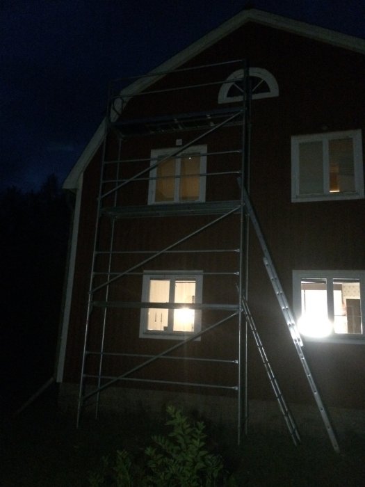 Byggställning vid ett rött hus i skymningen för att nå takets vindskiveplåt.