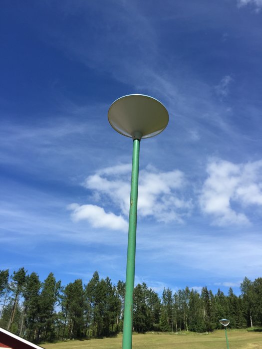 En utomhusstolpe med belysningsarmatur mot en klarblå himmel omgivet av träd och grönt gräs.