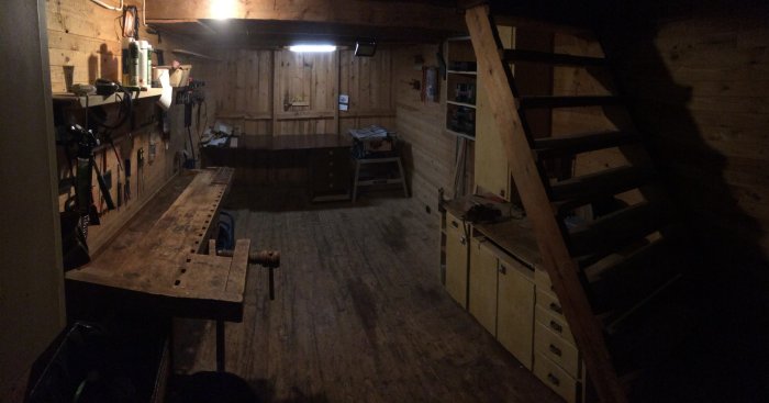 Gammaldags snickarbod med hyvelbänk, verktyg på väggen och trätrappa.