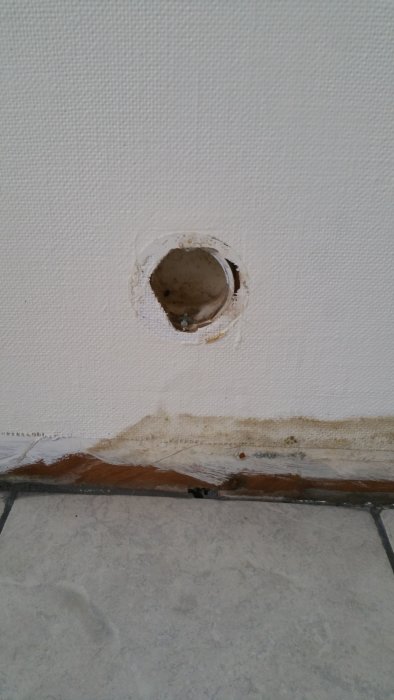 Vägg med gammal väv och ett hål för en tallriksventil, samt vattenskador längst ner vid golvet.