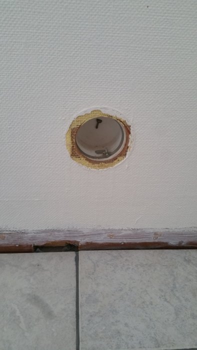 Öppning med gammal tallriksventil i en vägg klädd med texturerad tapet ovanför ett kaklat golv.