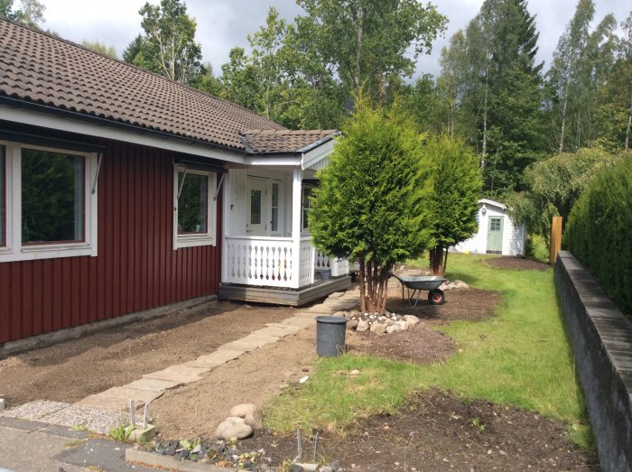 Framsidan av ett rött hus med vit veranda, gräsmatta, trädgårdsarbete pågår med grus och stenar.