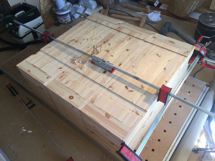 Träskiva balanserad med en tving för att kompensera för dess längre utstick på ett arbetsbord fyllt med verktyg och byggmaterial.