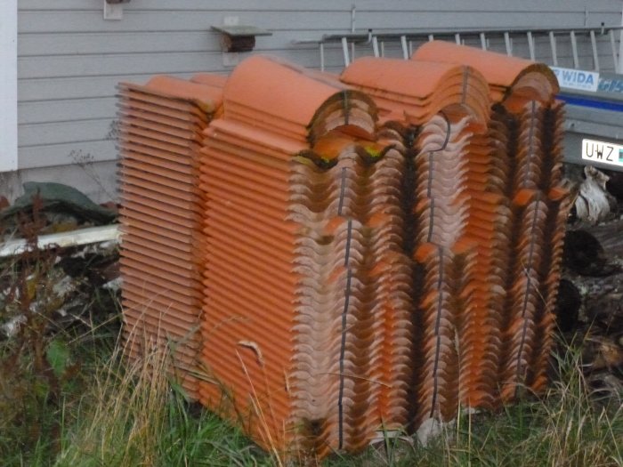 Stapel med över hundra orange betongtakpannor lagrade utomhus mot husvägg.