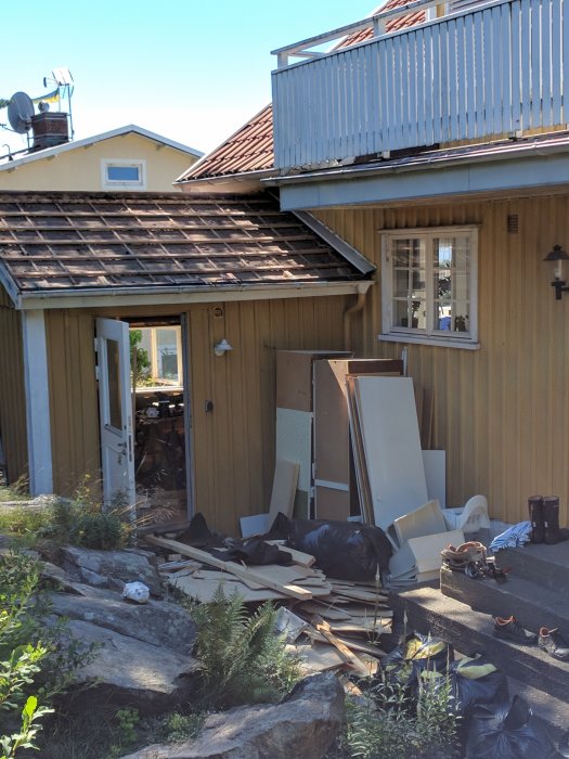 Ett trähus med skräp och byggmaterial utanför vid klippor och grönska, under klar himmel.