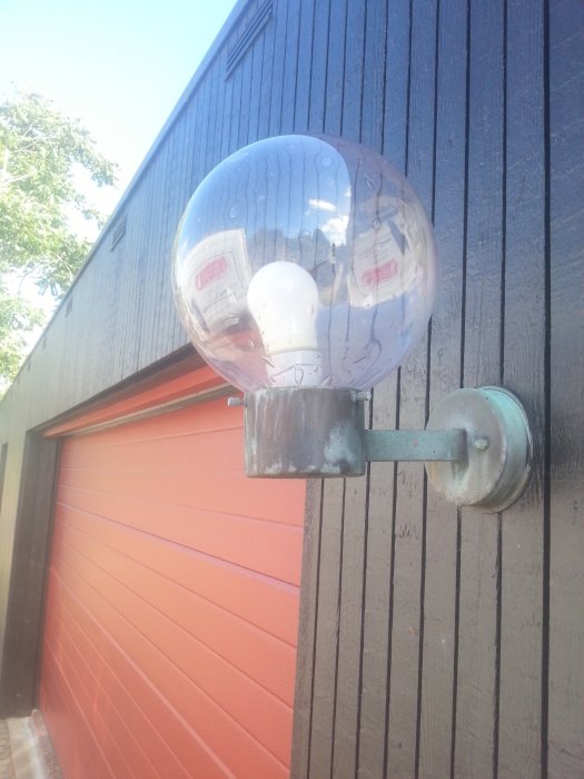 Stor rund utomhuslampa monterad på mörklaserad trävägg ovanför en orange garageport.