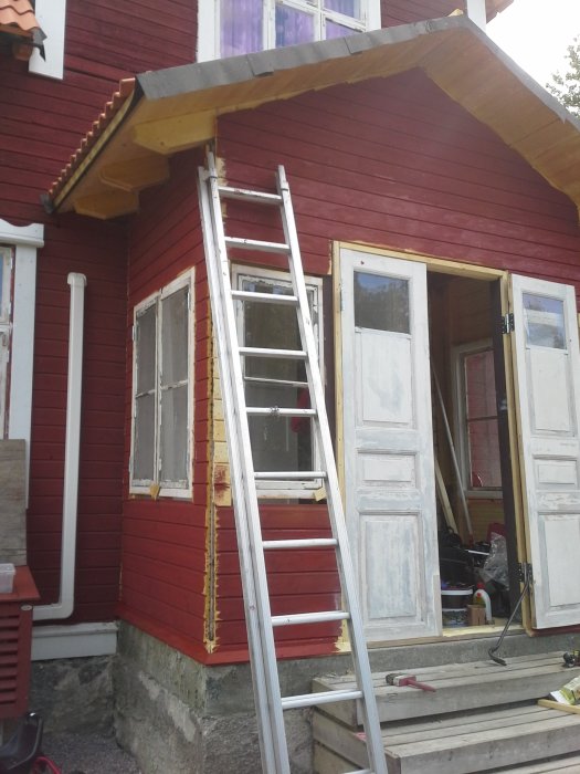 Renovering pågår på ett rött trähus, vita dörrar är borttagna och en stege står mot fasaden.