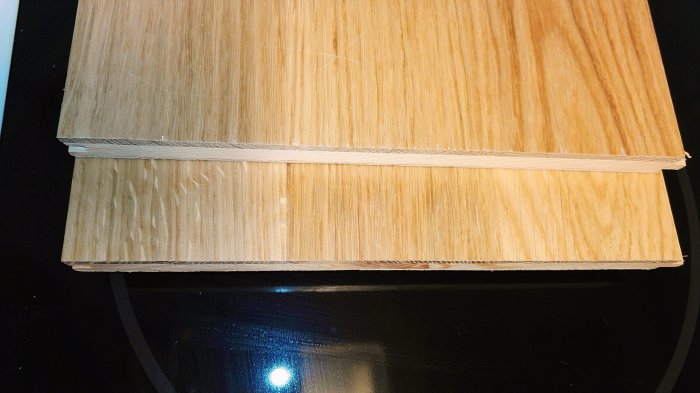 Två staplade plywoodskivor med synliga träfibrer och skikt, på en svart speglande yta.