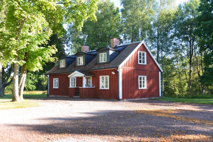 Röd trävilla med vita knutar och tre takkupor omgiven av gröna träd och grusgång.