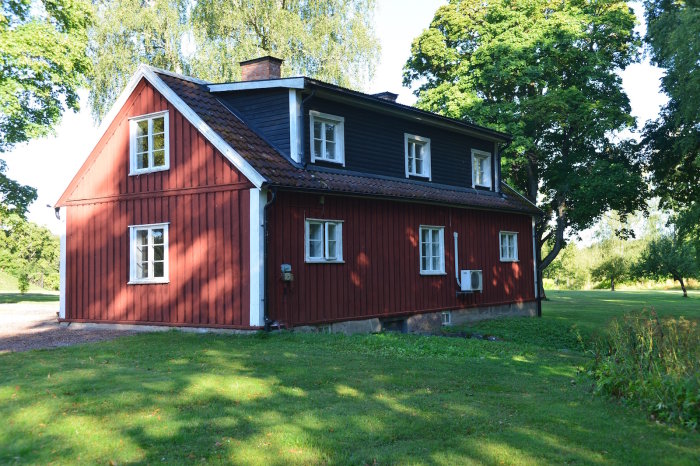 Rödmålad tvåvånings trävilla med svart tak och vita fönster, omgiven av grönt gräs och träd.