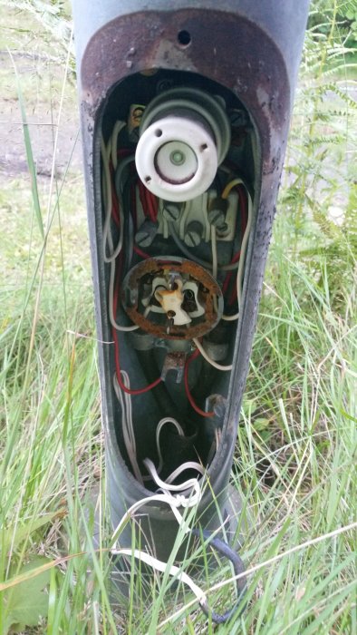 Öppet elektriskt uttag och kablar inom en metallstolpe för trädgårdsbelysning, med synlig rost och trassliga ledningar.