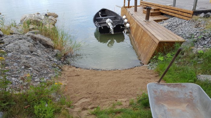 Altan vid vatten med en båt förtöjd och nyanlagd strand i förgrunden med skottkärra.