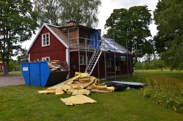 Hus i renoveringsfas med ställningar, delvis färdigställd takrivning och byggavfall som isolering och gamla takplåtar framför.