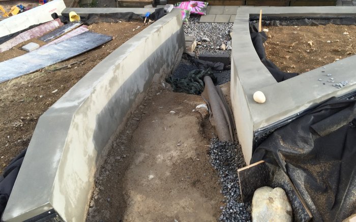 Pågående arbete med grundläggning och stödmur i en trädgård, inklusive markduk och dräneringsgrus.