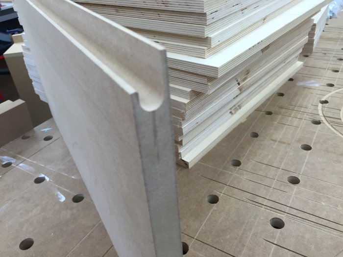 Björkplywoodskiva med fräst fingergrepp står framför en stapel plywood, verktygshål i arbetsbänk syns.