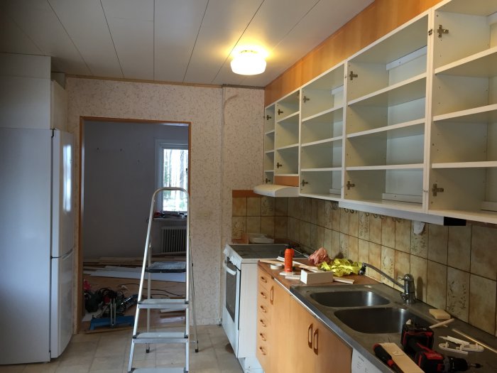 Ett kök under renovering med borttagna skåpluckor, en stege och renoveringsmaterial synliga.