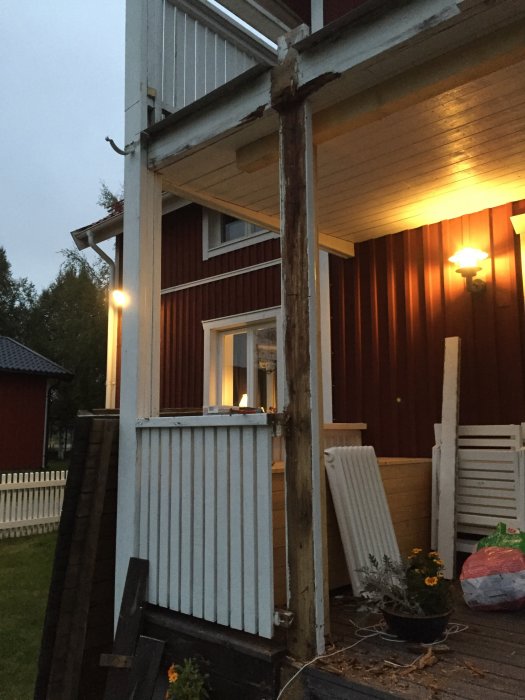 Genomrutten trästolpe stödjer en balkong med synliga skador, bredvid ett rött hus på kvällen.