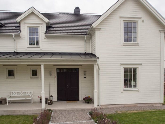 Ett vitt enplanshus med svart falsat plåttak, veranda och en garageingång.