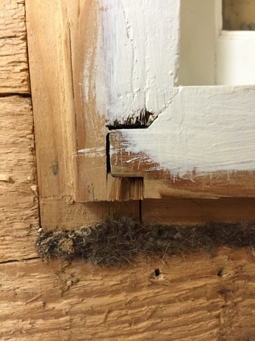 Sliten vitmålad fönsterkarm från 1945 med spricka och skador intill karmens hörn mot träväggen.