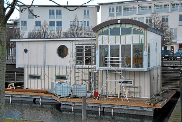 Hus på flotte med vattenmagasin under konstruktion, omgivet av vatten och andra byggnader.