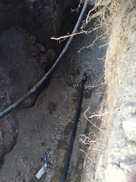 Grävd grop med nytt svart vattenrör och gamla rotrester i jorden efter ledningsbytet.