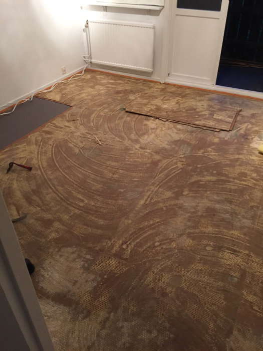 Delvis avtäckt laminatgolv visar underliggande masonite och gamla plank i ett rum under renovering.