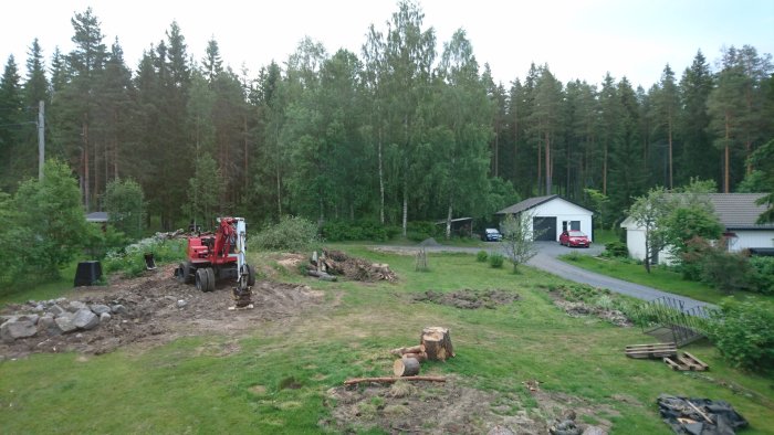 En trädgård med en sönder trasig grävmaskin på tre hjul, omgivande träd och en bostad i bakgrunden.