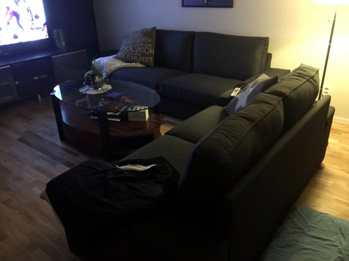 Vardagsrum med svart soffa, glasbord och tänd TV, mjuka textilier på soffan och ett dekorativt kuddfodral med text.